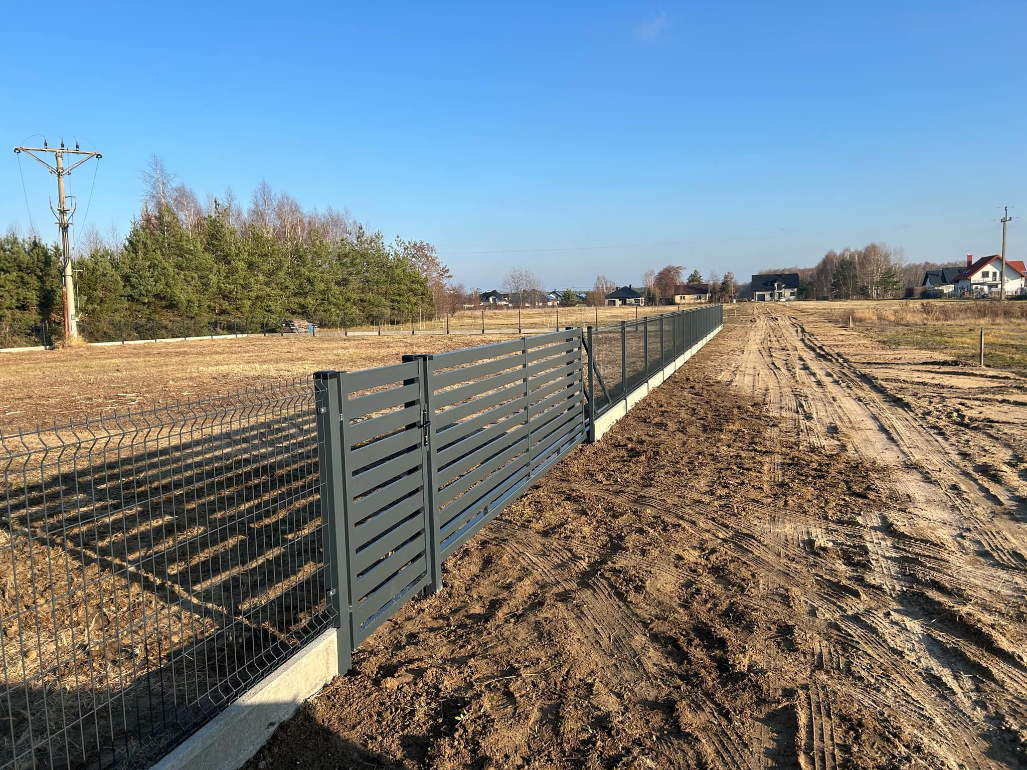 Długi, szary płot panelowy z betonową podmurówką, częściowo z siatki, na nierównym terenie z widocznymi śladami opon, w tle domy i drzewa pod błękitnym niebem.