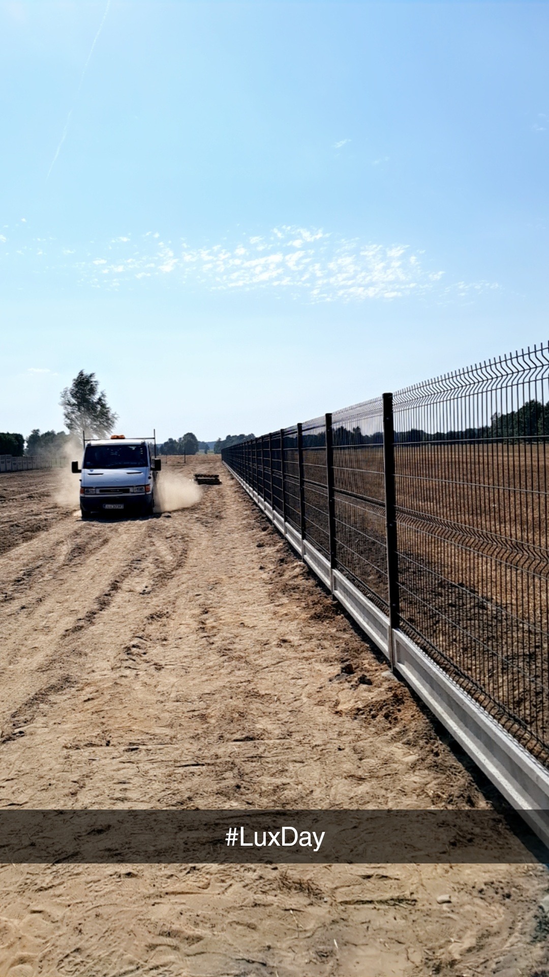 Długi, czarny panelowy płot z betonową podmurówką, ciągnący się wzdłuż polnej drogi, po której jedzie biały dostawczy samochód wzbijający tumany kurzu.