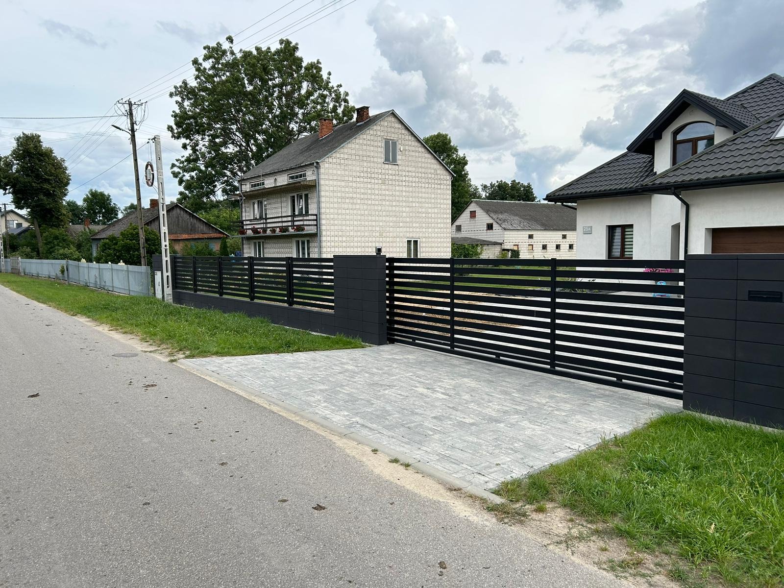 Nowoczesna, pozioma brama wjazdowa i przęsła ogrodzeniowe w kolorze antracytowym, z szarej kostki brukowej podjazd, w tle domy jednorodzinne.