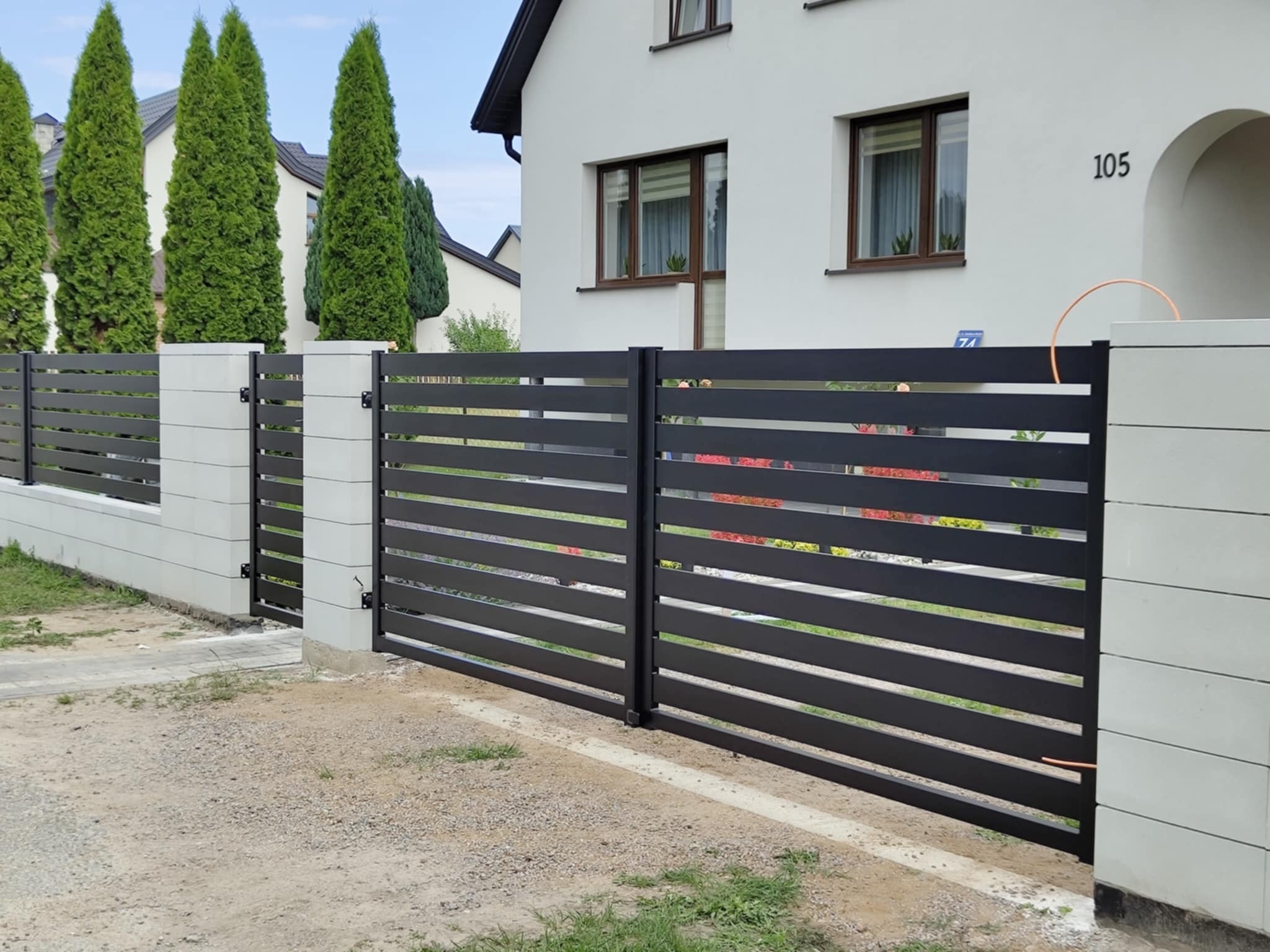 Nowoczesna, pozioma brama wjazdowa z czarnych paneli, osadzona na jasnych, betonowych słupkach. W tle biały dom z numerem 105 i zielone tuje.