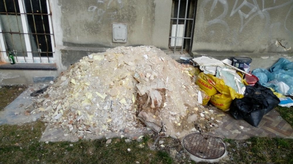 Sterta gruzu i odpadów budowlanych przed budynkiem z oknami w kratach, obok leżą worki i puszki po farbach, a w tle widoczny jest fragment trawnika i studzienka kanalizacyjna.