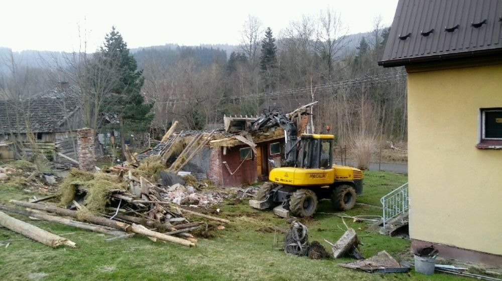 Żółta maszyna budowlana Mecalac demontuje częściowo zburzony budynek z czerwonej cegły i drewna na zarośniętej trawie, w tle widoczne drzewa i fragment budynku mieszkalnego.