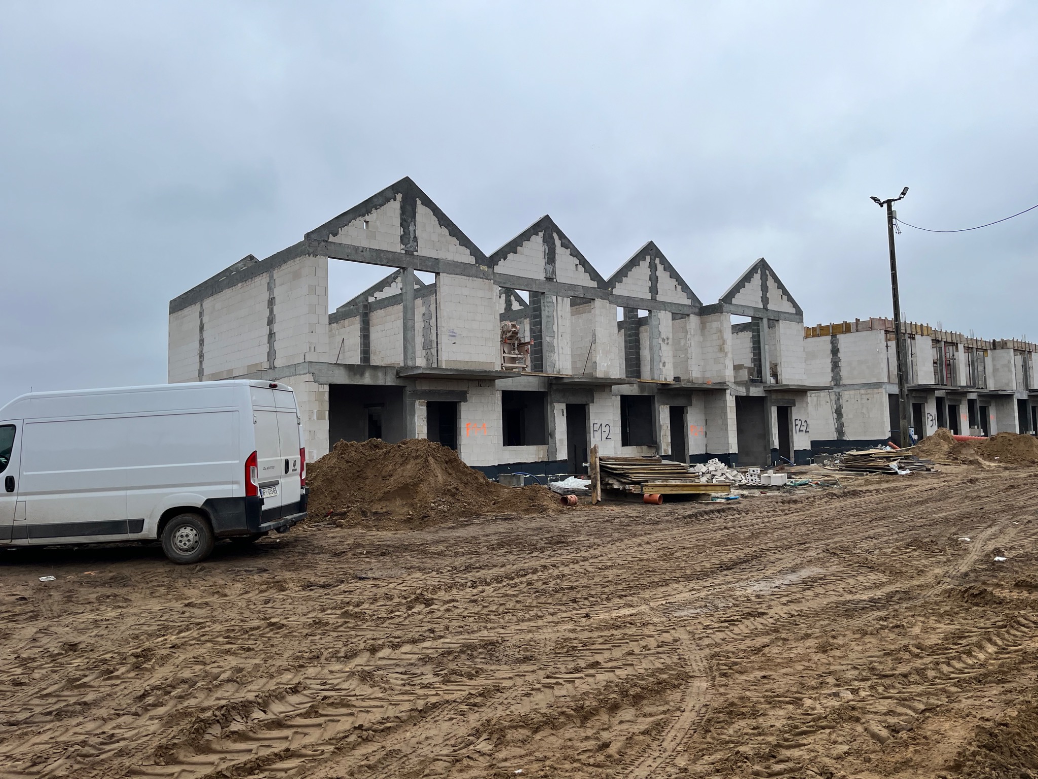 Surowe, niedokończone budynki szeregowe z białych bloczków, widoczne ślady zaprawy, na pierwszym planie biały samochód dostawczy i błotniste podłoże z wyraźnymi śladami opon.