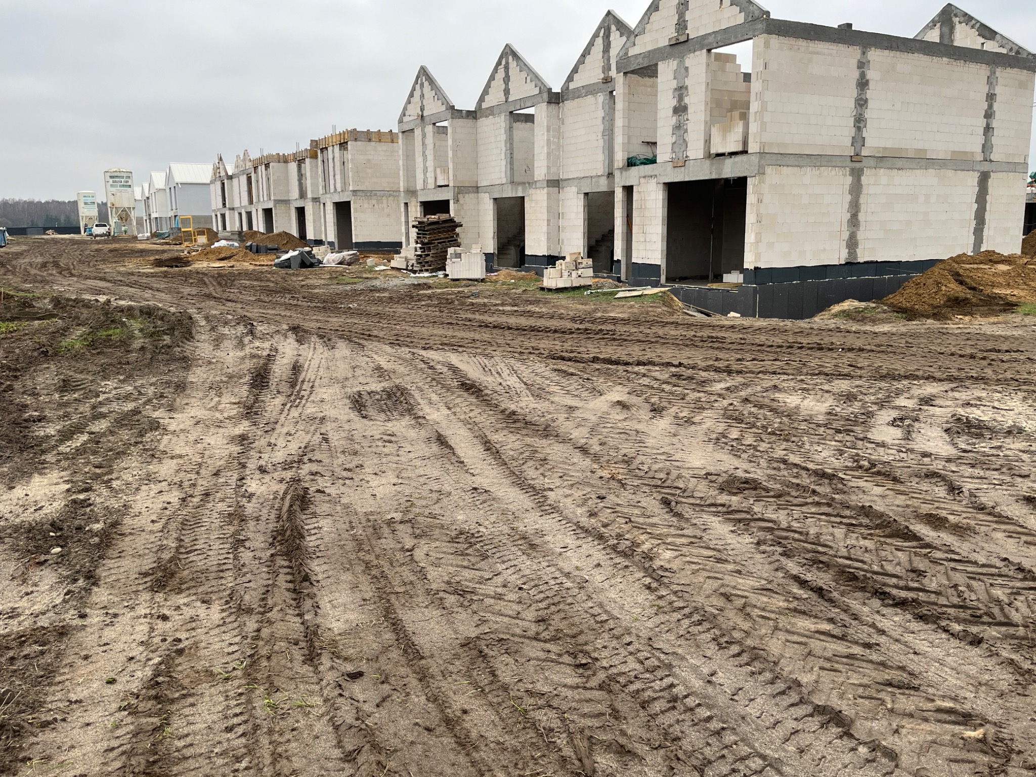 Wczesny etap budowy szeregu domów jednorodzinnych z białego bloczka, widoczne fundamenty z izolacją, w tle silosy na cement, na pierwszym planie błotnista droga z wyraźnymi śladami opon.