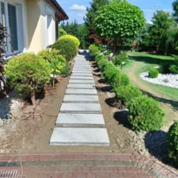 Ścieżka z szarych płyt betonowych prowadząca przez zadbany ogr&oacute;d z krzewami i drzewami, widok w kierunku domu z czerwonej kostki brukowej.