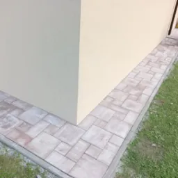 Ścieżka z kostki brukowej ułożona wzdłuż budynku z beżową elewacją, widoczna krawędź trawnika i fragment zagęszczarki w tle.