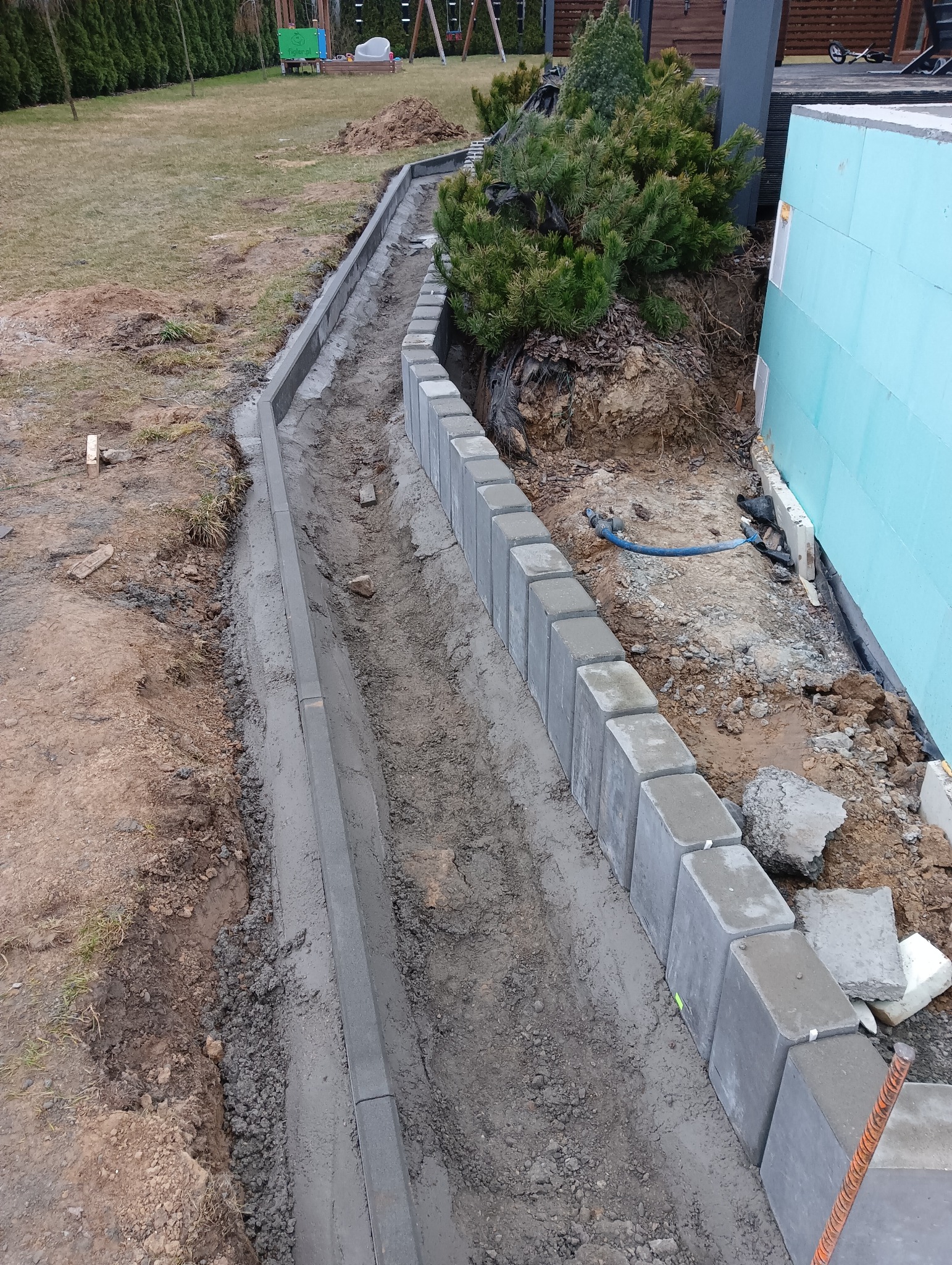 Świeżo ułożony obrzeże z betonowych kostek oddzielające trawnik od ścieżki, widoczny fragment budynku z izolacją, ziemia wokół w trakcie prac ziemnych.