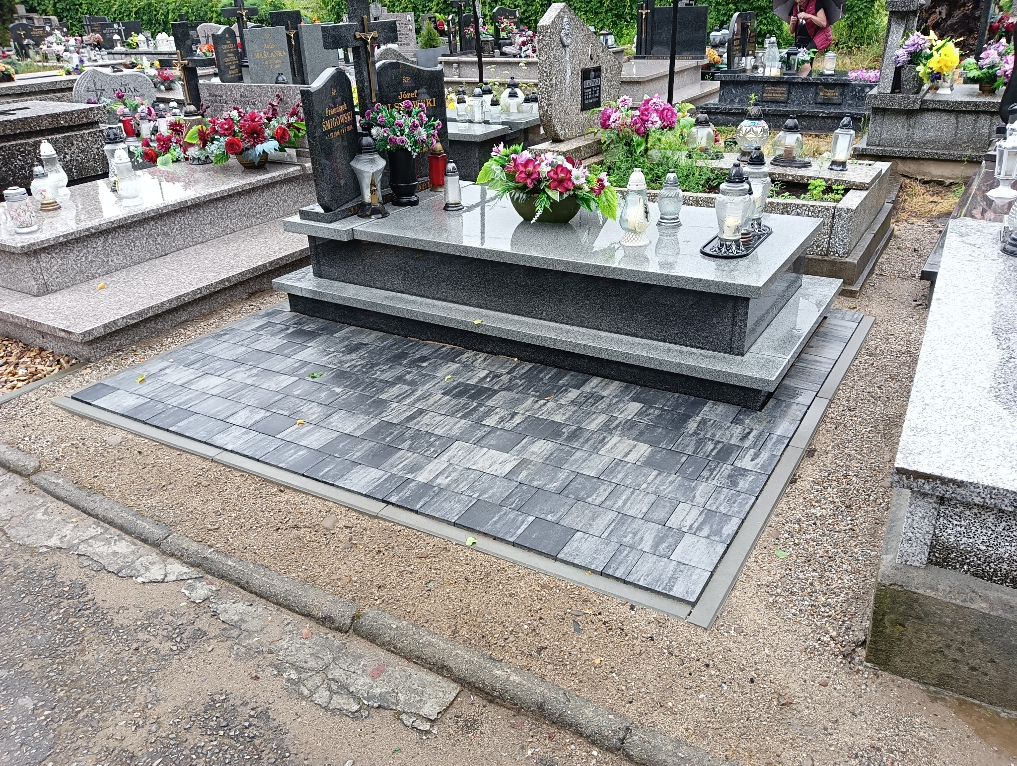 Częściowo widoczny nagrobek z szarego kamienia z kwiatami i zniczami, otoczony nową, szarą kostką brukową ułożoną na cmentarzu, widok z poziomu gruntu.