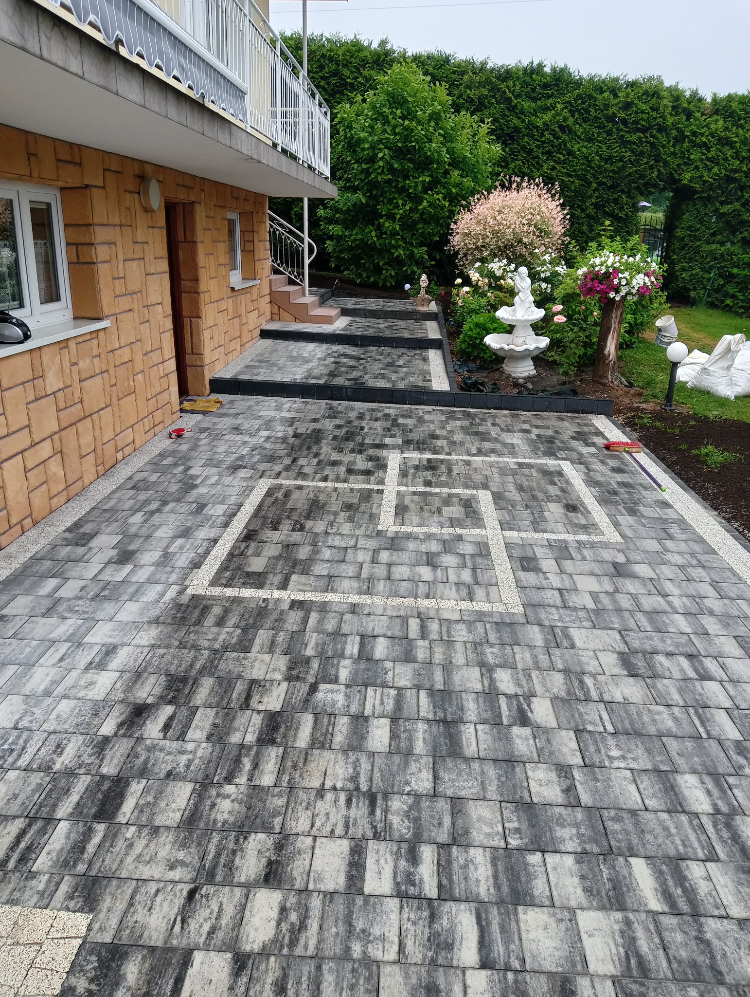 Ułożona kostka brukowa w szaro-białej kolorystyce z geometrycznym wzorem, widoczny fragment domu z kamienną elewacją i ogród z fontanną w tle.