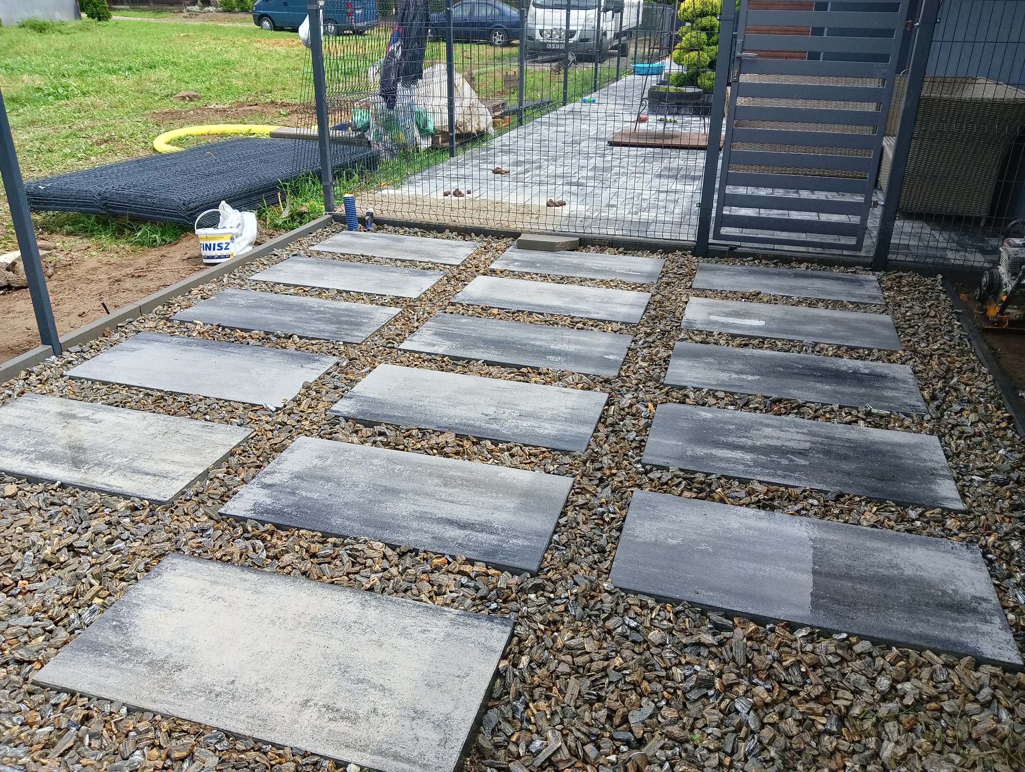 Ścieżka z prostokątnych płyt betonowych ułożonych na żwirze, otoczona siatką ogrodzeniową, widoczny przygotowany materiał do montażu ogrodzenia i narzędzia.