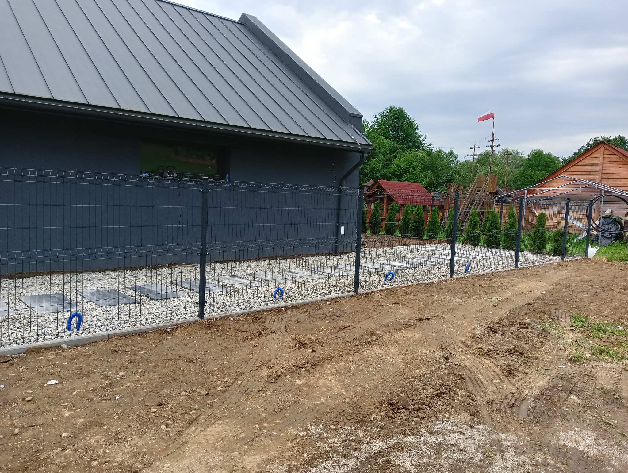 Nowoczesne ogrodzenie panelowe w kolorze grafitowym, z betonową podmurówką i podłożem z białego żwiru, uzupełnione niebieskimi elementami mocującymi, na tle domu z szarą blachodachówką i częściowo...