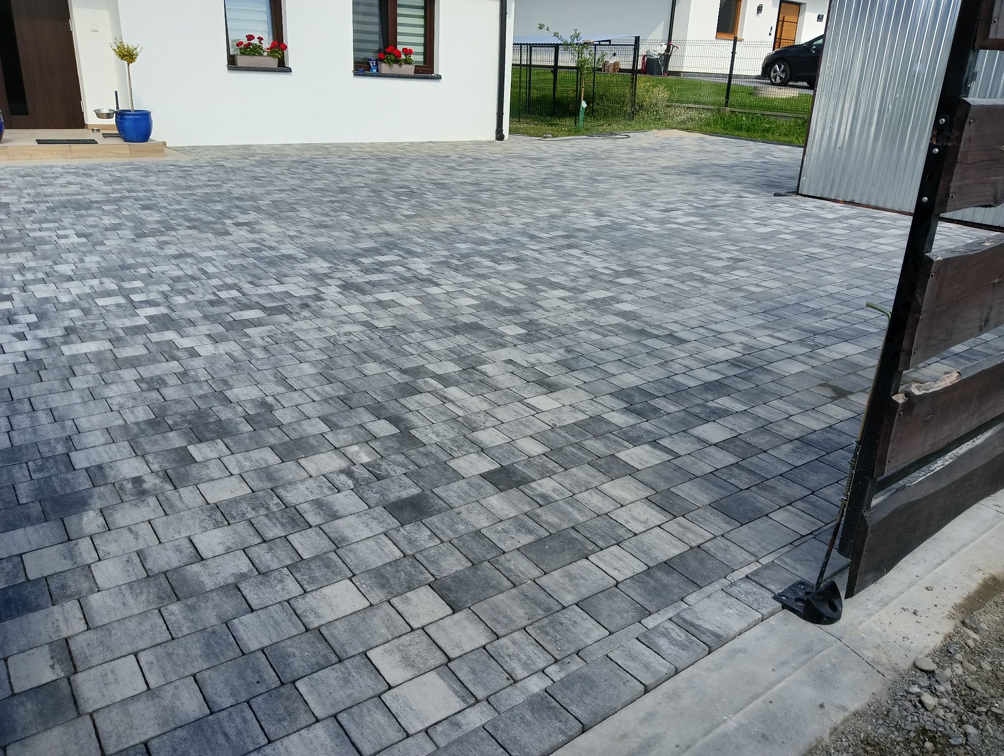 Ułożona szara kostka brukowa na podjeździe przed domem z białymi ścianami i czerwonymi kwiatami w oknach, widok z perspektywy lekko z boku, fragment ogrodzenia po prawej stronie.