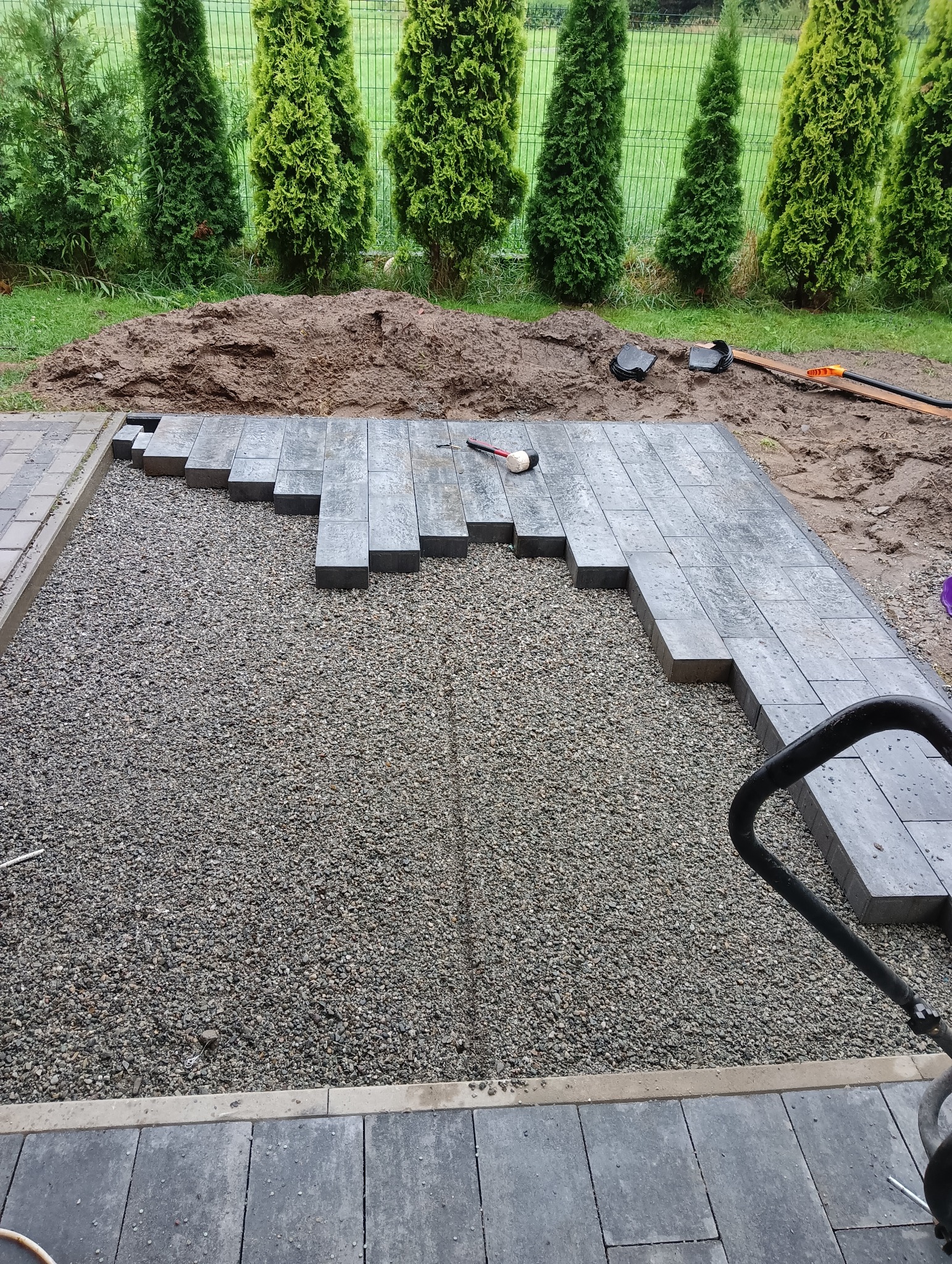 Szare, prostokątne płyty chodnikowe układane na podsypce żwirowej, z widocznym gumowym młotkiem i ubijarką w tle, na obrzeżach trawnika z tujami.