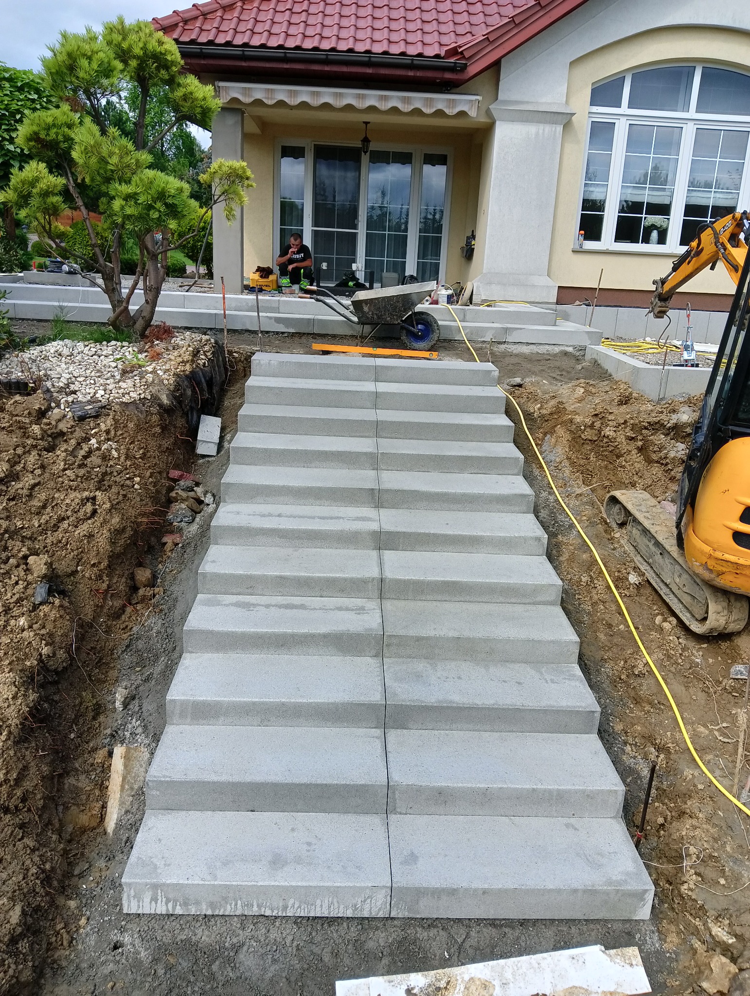 Montaż schodów kamiennych z jasnoszarego betonu prefabrykowanego, widok z dołu na gotowe stopnie, obok wykop ziemny z widocznymi korzeniami, minikoparka i pracownik w tle.