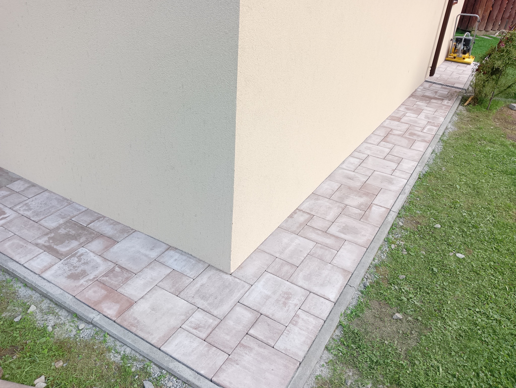 Ścieżka z kostki brukowej ułożona wzdłuż budynku z beżową elewacją, widoczna krawędź trawnika i fragment zagęszczarki w tle.