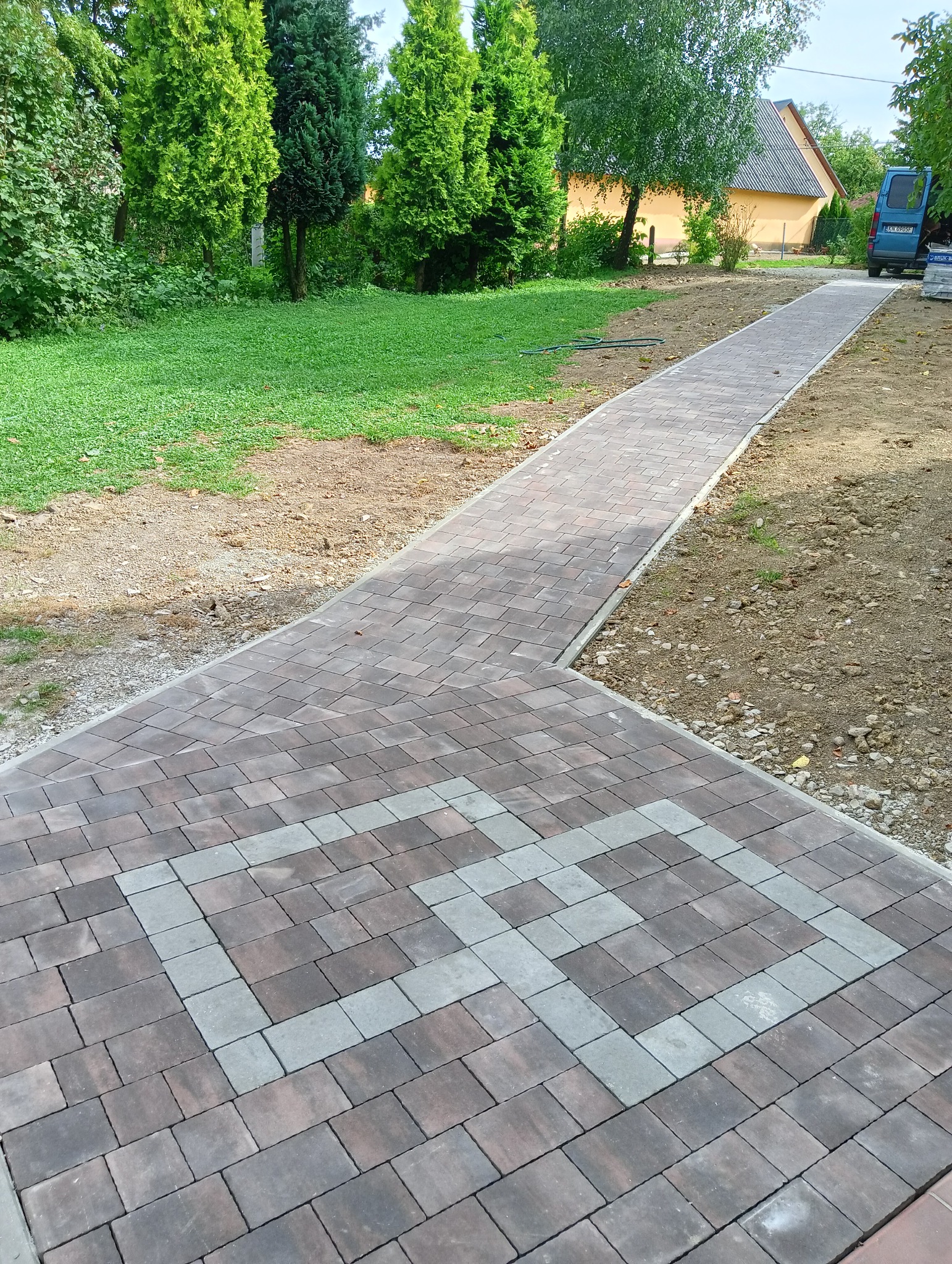 Nowo ułożona ścieżka z kostki brukowej w Nowym Sączu, z dekoracyjnym wzorem z jasnych i ciemnych odcieni, prowadząca przez trawnik w kierunku domu i zaparkowanego samochodu.
