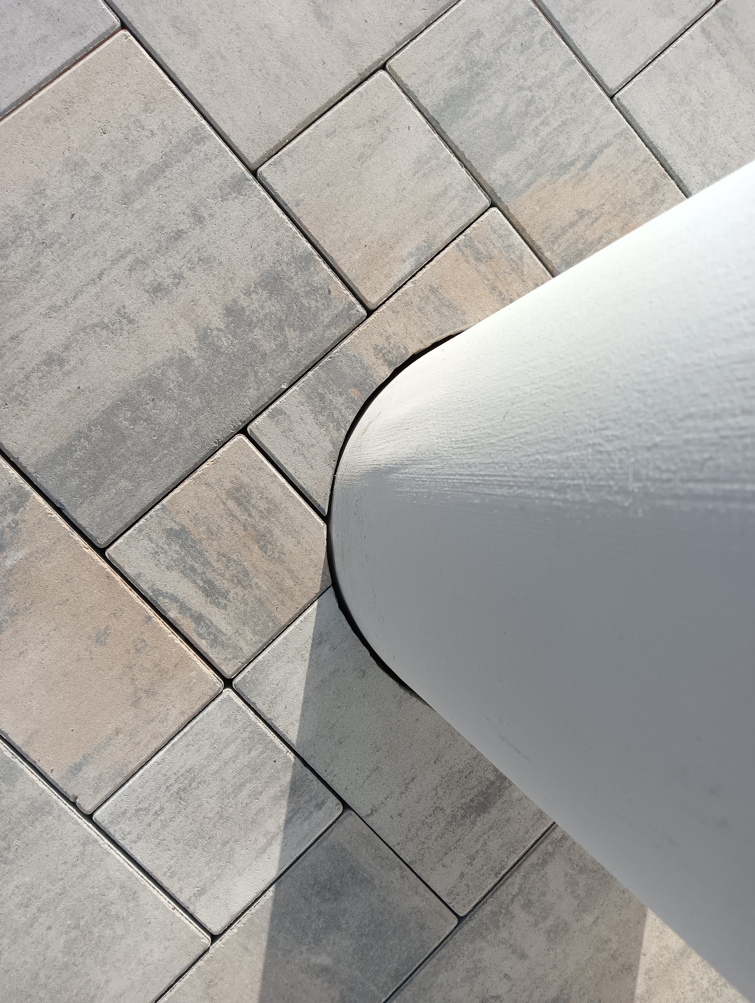 Szare, kwadratowe kostki brukowe ułożone wokół białej, okrągłej kolumny, widoczne z góry, z delikatnym cieniem.