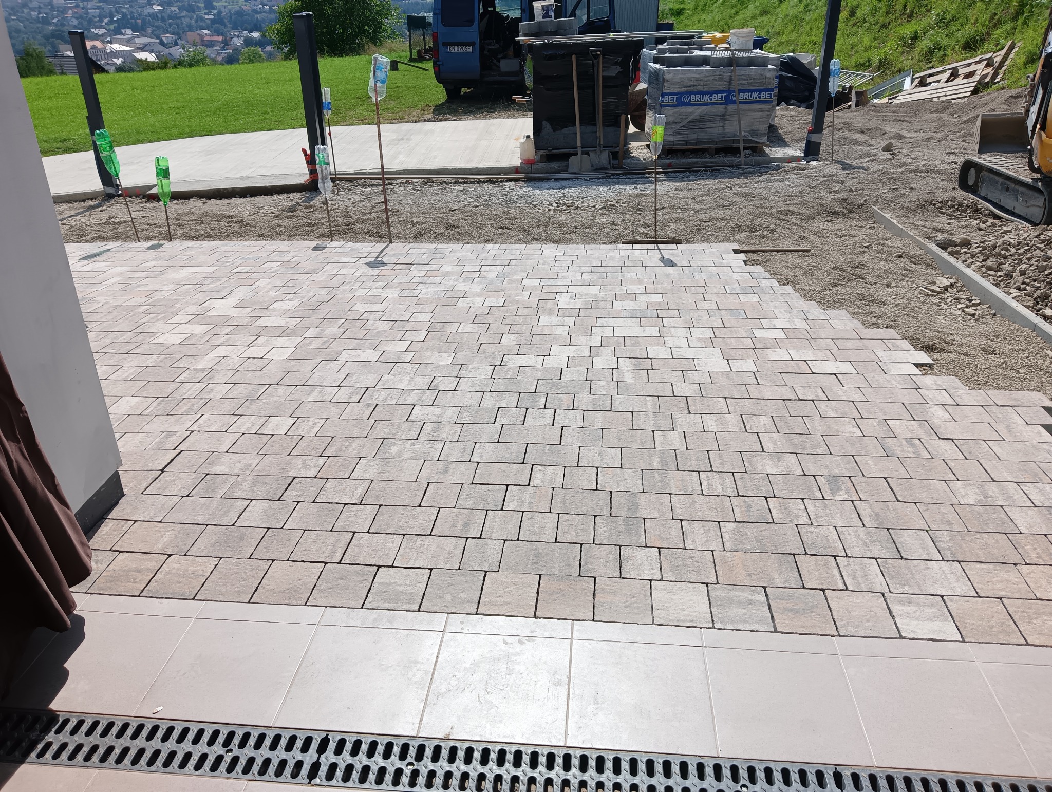 Fragment nowej nawierzchni z kostki brukowej w odcieniach szarości, układanej w jodełkę, z widocznym odwodnieniem liniowym, przygotowanym podłożem i materiałami budowlanymi w tle, w słoneczny dzień.