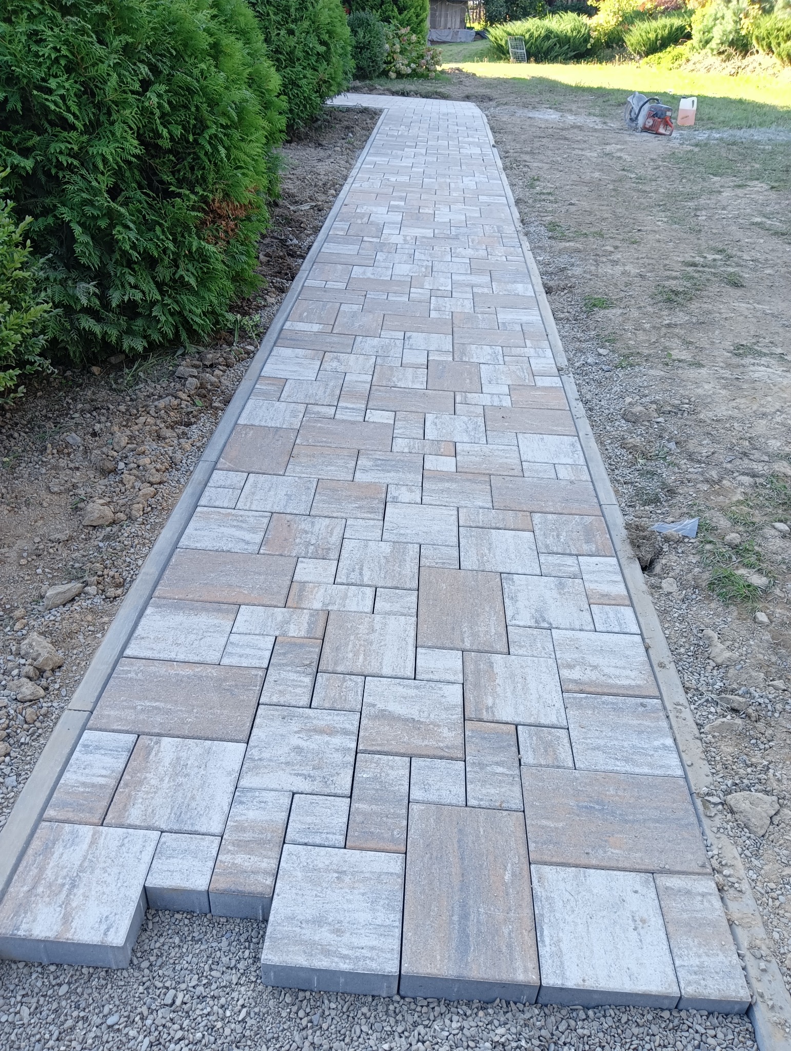 Ścieżka z nieregularnych, szaro-beżowych kostek brukowych w trakcie układania, obrzeża betonowe, widoczna piła do cięcia kostki w tle.