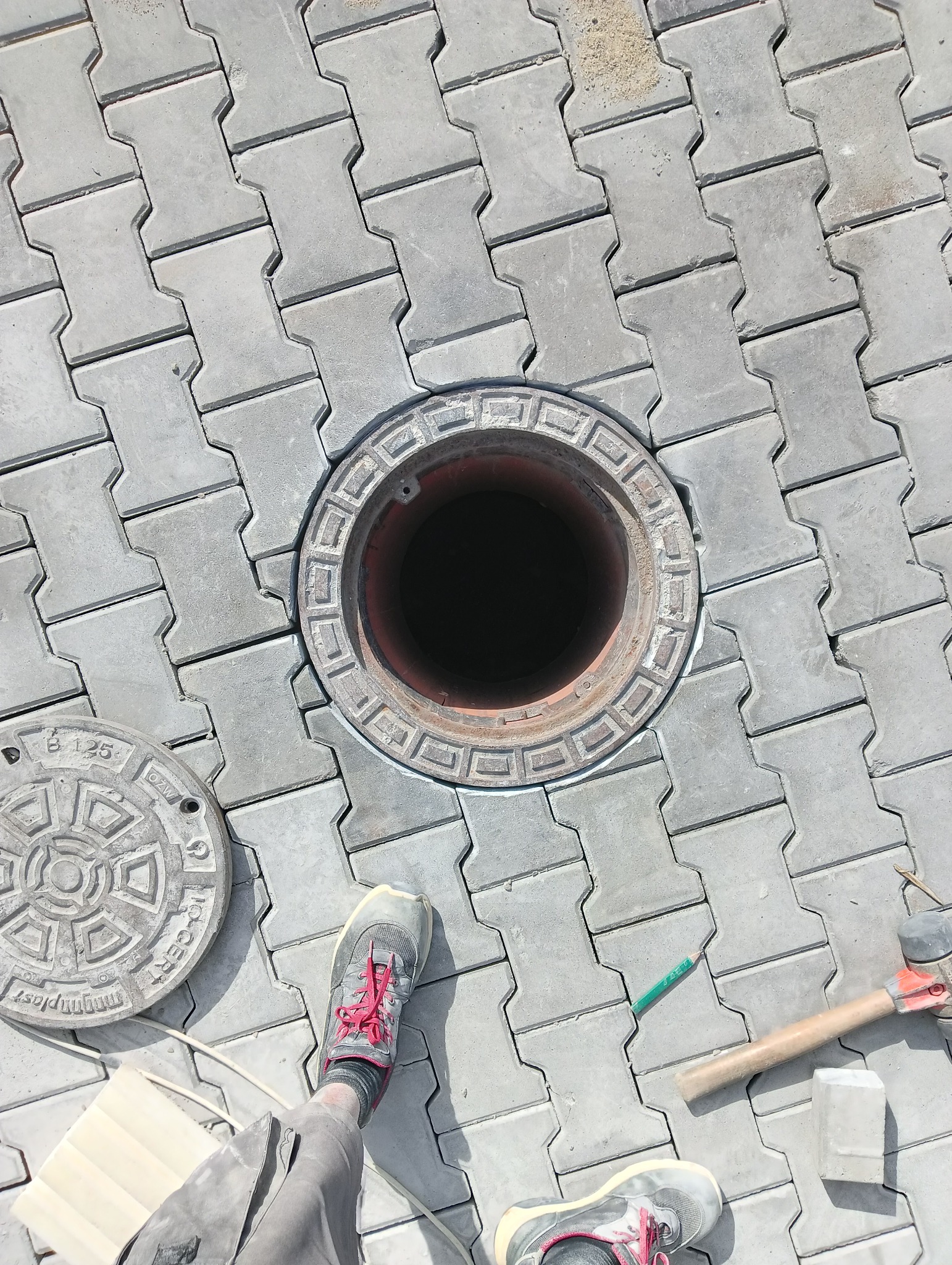 Układanie szarej kostki brukowej typu 'I' wokół studzienki kanalizacyjnej z widoczną otwartą pokrywą i narzędziami, w trakcie prac wykończeniowych.