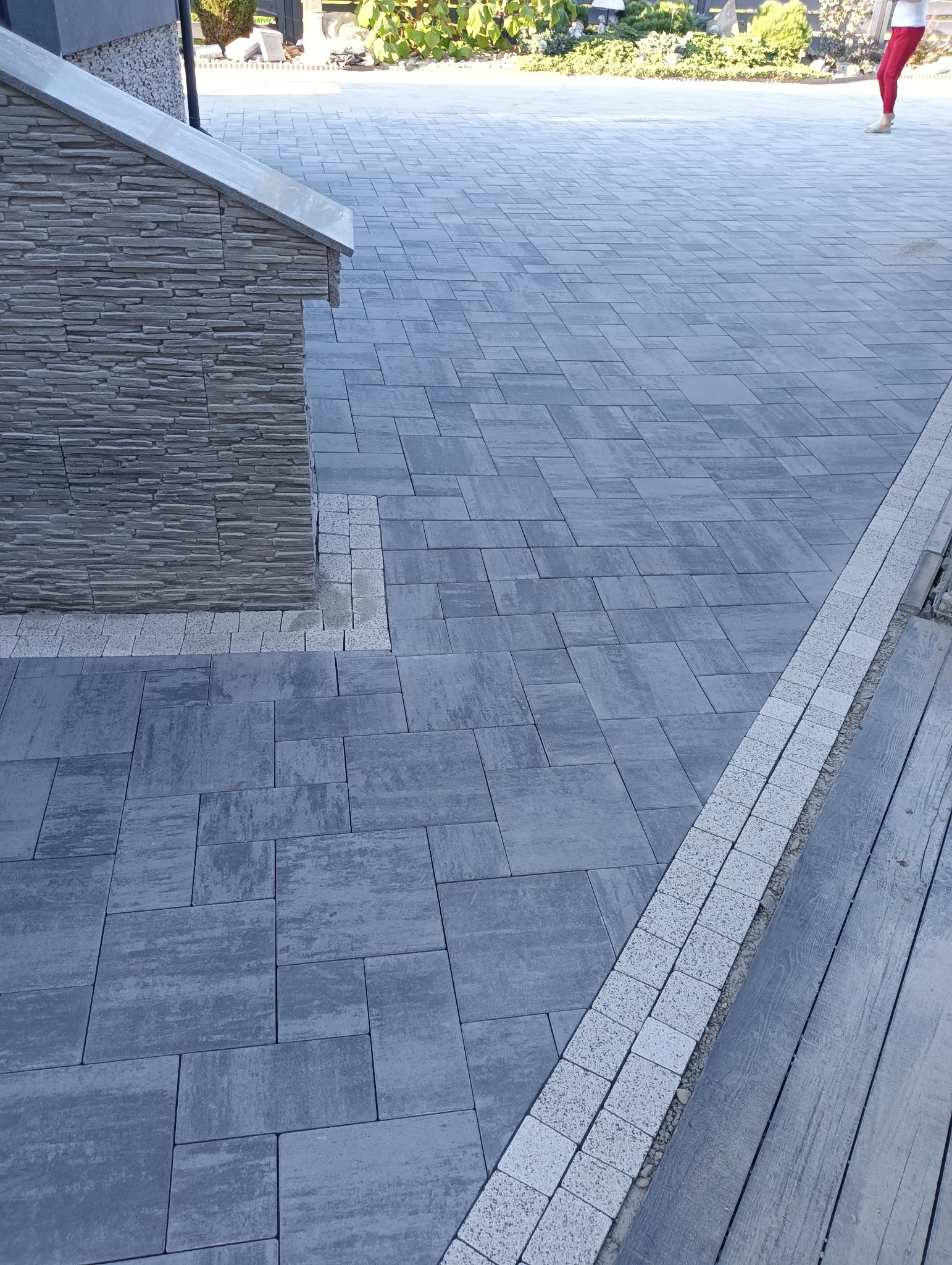 Szara kostka brukowa ułożona na podjeździe, z obrzeżem z jasnej kostki, przylegająca do ściany budynku wykończonej kamieniem elewacyjnym.