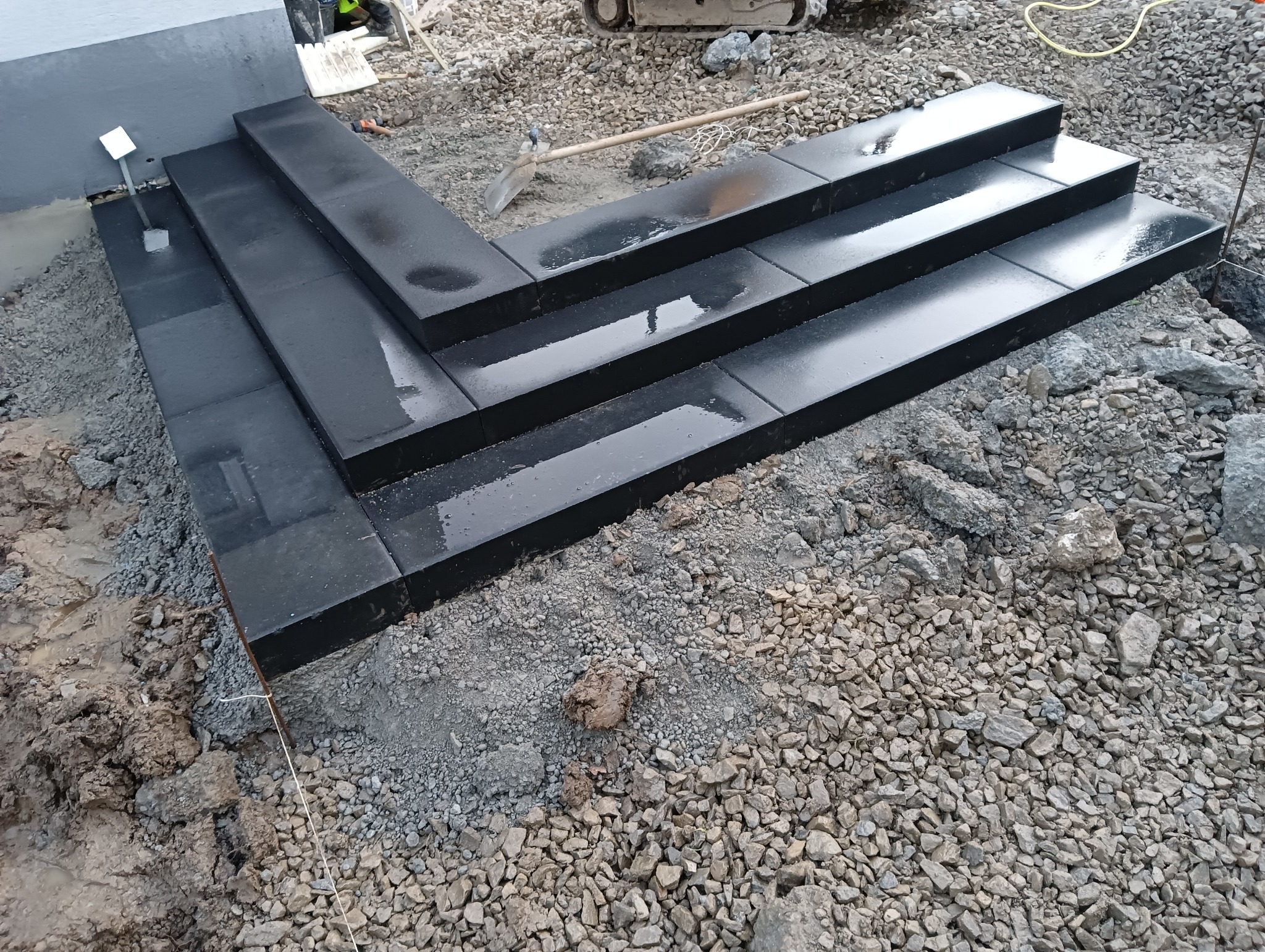 Trzy stopnie schodów z ciemnej, mokrej kostki ułożone na żwirowym podłożu, z widoczną łopatą i fragmentem mini-koparki w tle.