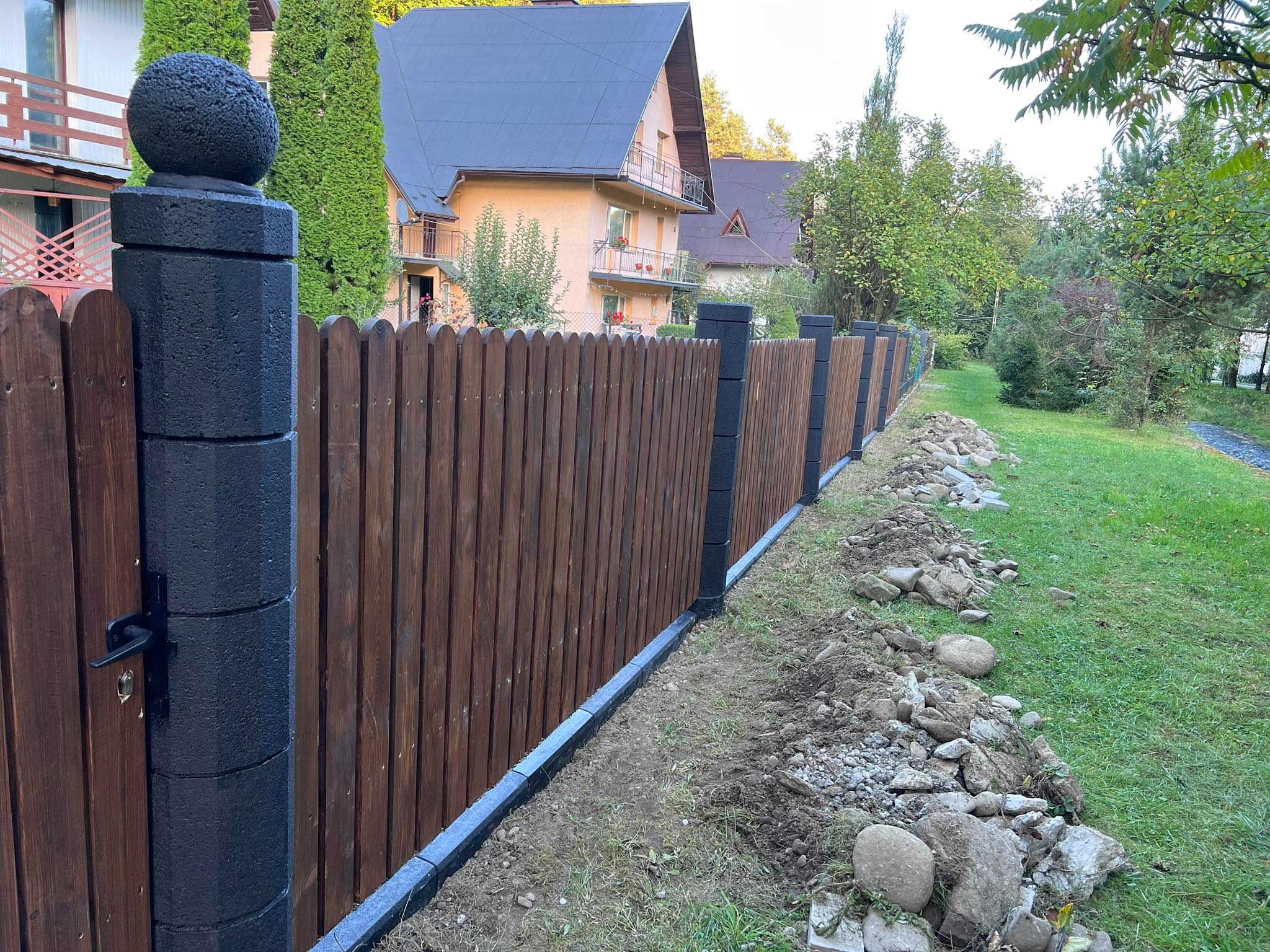 Nowo postawione drewniane ogrodzenie z czarnymi, betonowymi słupkami zwieńczonymi kulami, obrzeża z kostki betonowej, teren po pracach ziemnych z kamieniami.