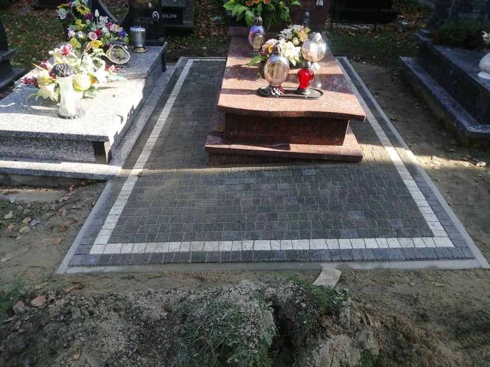 Ciemnoszara kostka brukowa ułożona wokół nagrobka z jasną obwódką, widoczne kwiaty i znicze na nagrobkach w tle.