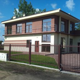 Tani Dom - Dwukondygnacyjny dom jednorodzinny z płaskim dachem, brązowo-beżową elewacją, balkonem i metalowym ogrodzeniem z bramą przesuwną, otoczony trawnikiem i zielenią.