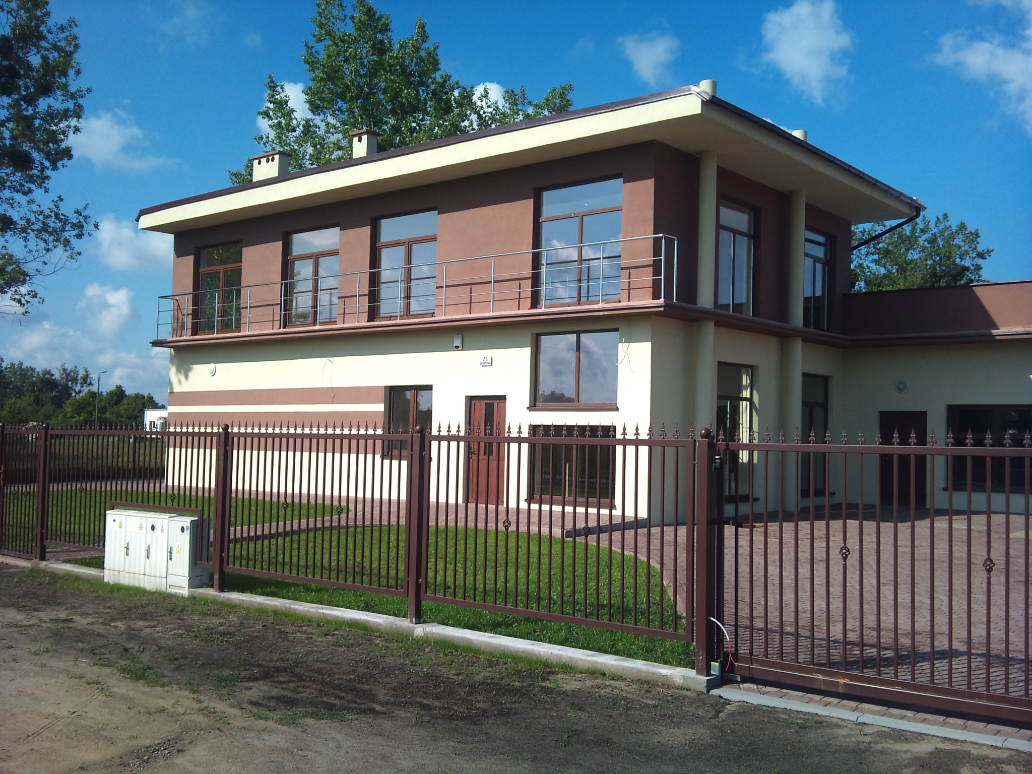 Dwukondygnacyjny dom jednorodzinny z płaskim dachem, brązowo-beżową elewacją, balkonem i metalowym ogrodzeniem z bramą przesuwną, otoczony trawnikiem i zielenią.