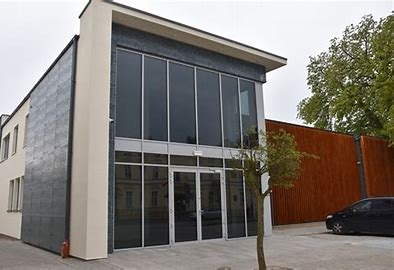 Nowoczesny budynek usługowy z dużymi oknami i elewacją z ciemnej płytki, drzewko na pierwszym planie, obok drewniane ogrodzenie