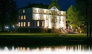 Elegancki, podświetlony budynek dwukondygnacyjny z licznymi oknami łukowymi, widziany nocą z perspektywy lustra wodnego, z zielonym trawnikiem i drzewami w tle.