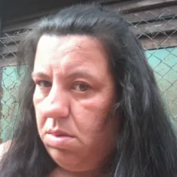 Portret kobiety z ciemnymi włosami na tle drewnianej ściany i siatki.