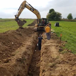 Żółta koparka CAT pogłębia wykop w ziemi na tle zielonego pola, w wykopie i obok niego stoją pracownicy, a w tle widoczny jest samochód z otwartym bagażnikiem i rury.