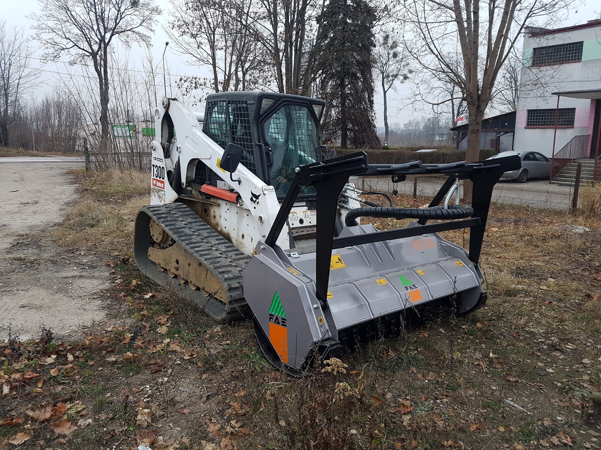 Biała minikoparka gąsienicowa Bobcat T300 z frezem do pni FAE, stojąca na ziemi pokrytej liśćmi, na tle drzew i budynku.