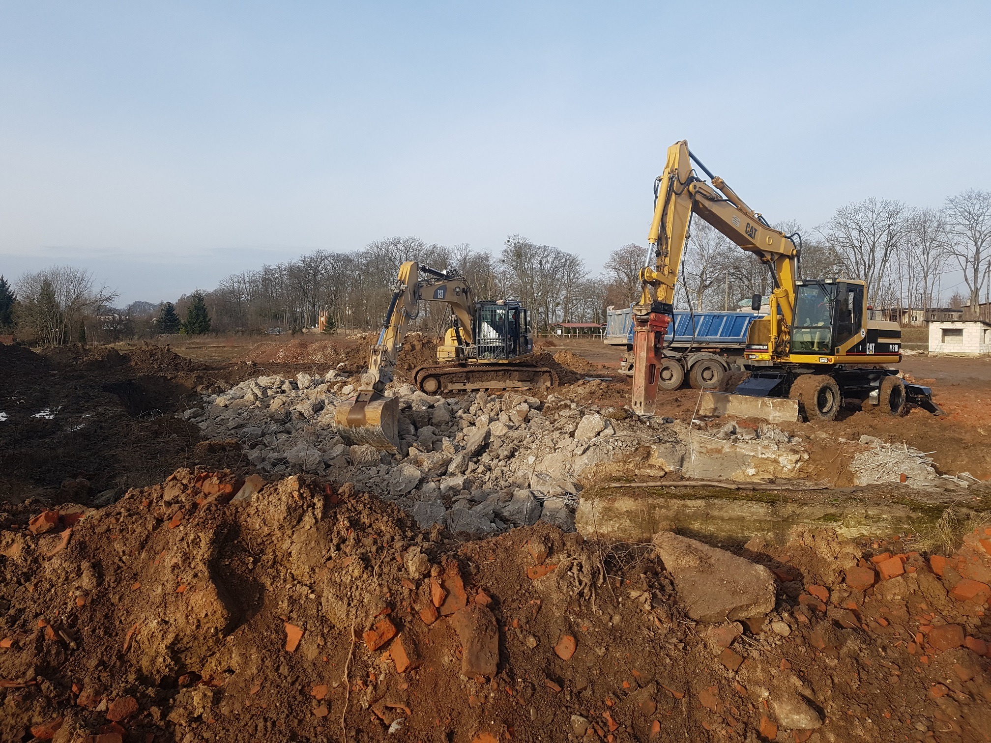 Dwa żółte koparki CAT na placu budowy, jedna z młotem hydraulicznym i wywrotką, gruz i cegły w tle, drzewa bez liści i błękitne niebo.