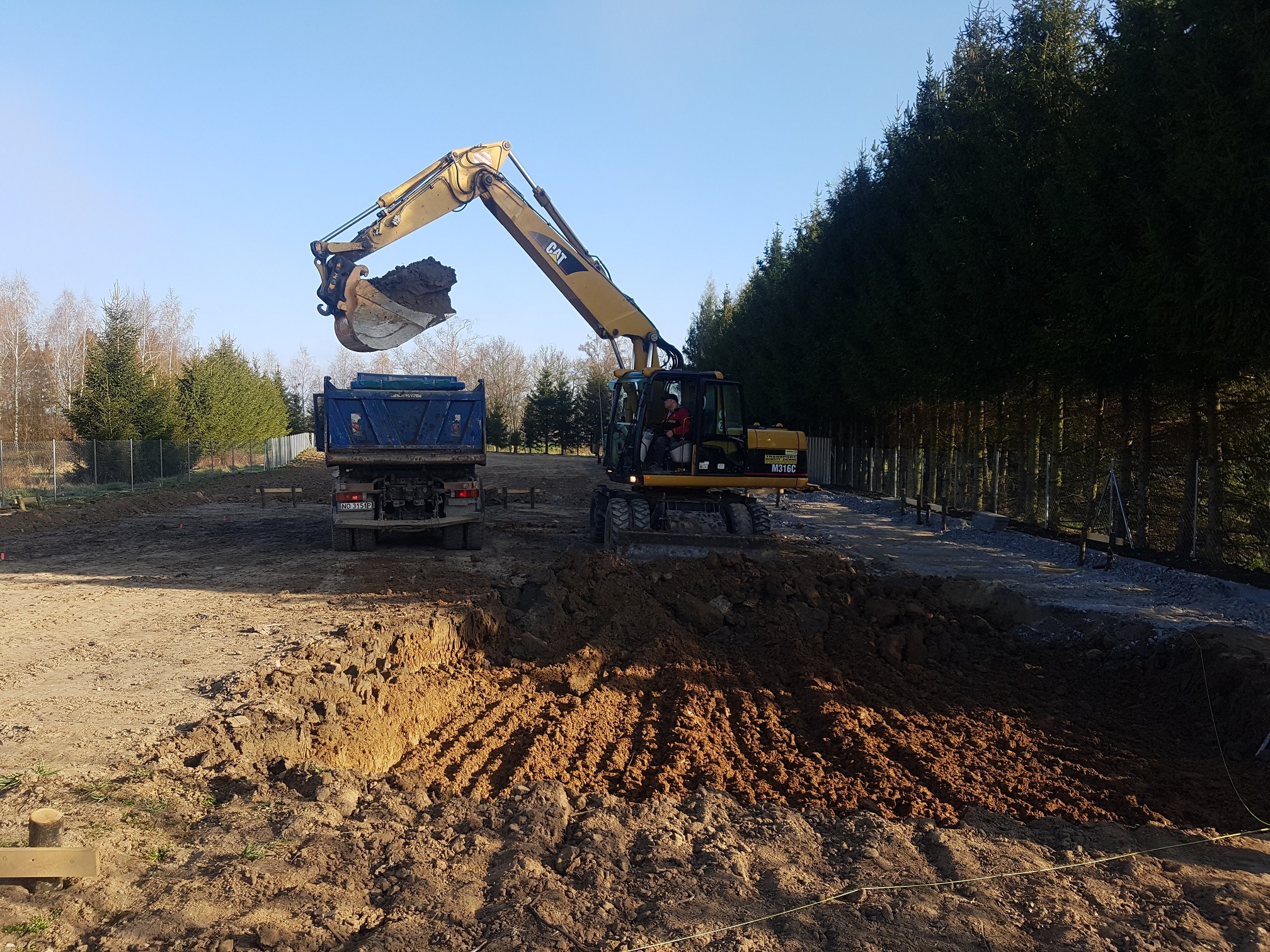 Żółta koparka CAT M316C ładuje ziemię do niebieskiej wywrotki na tle rzędu ciemnozielonych drzew iglastych i błękitnego nieba.