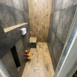 Wnętrze kabiny prysznicowej w trakcie prac wykończeniowych, z widocznymi płytkami imitującymi drewno na podłodze i ścianach, instalacją wodną z pomarańczowymi elementami oraz półką wbudowaną w ścianę.