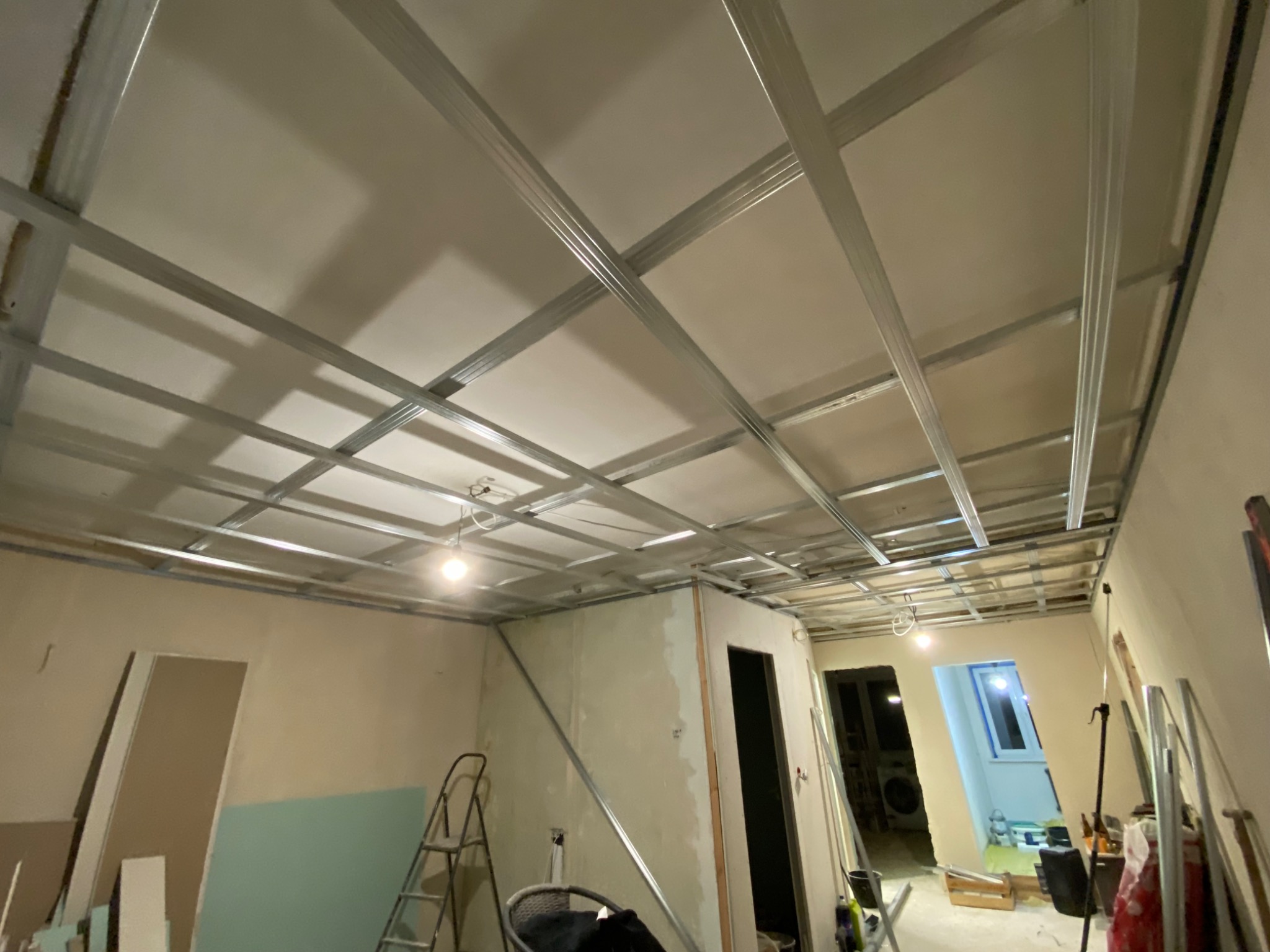Metalowa konstrukcja sufitu podwieszanego z płyt karton gips, widoczne profile i fragmenty ścian w trakcie remontu, drabina w tle.