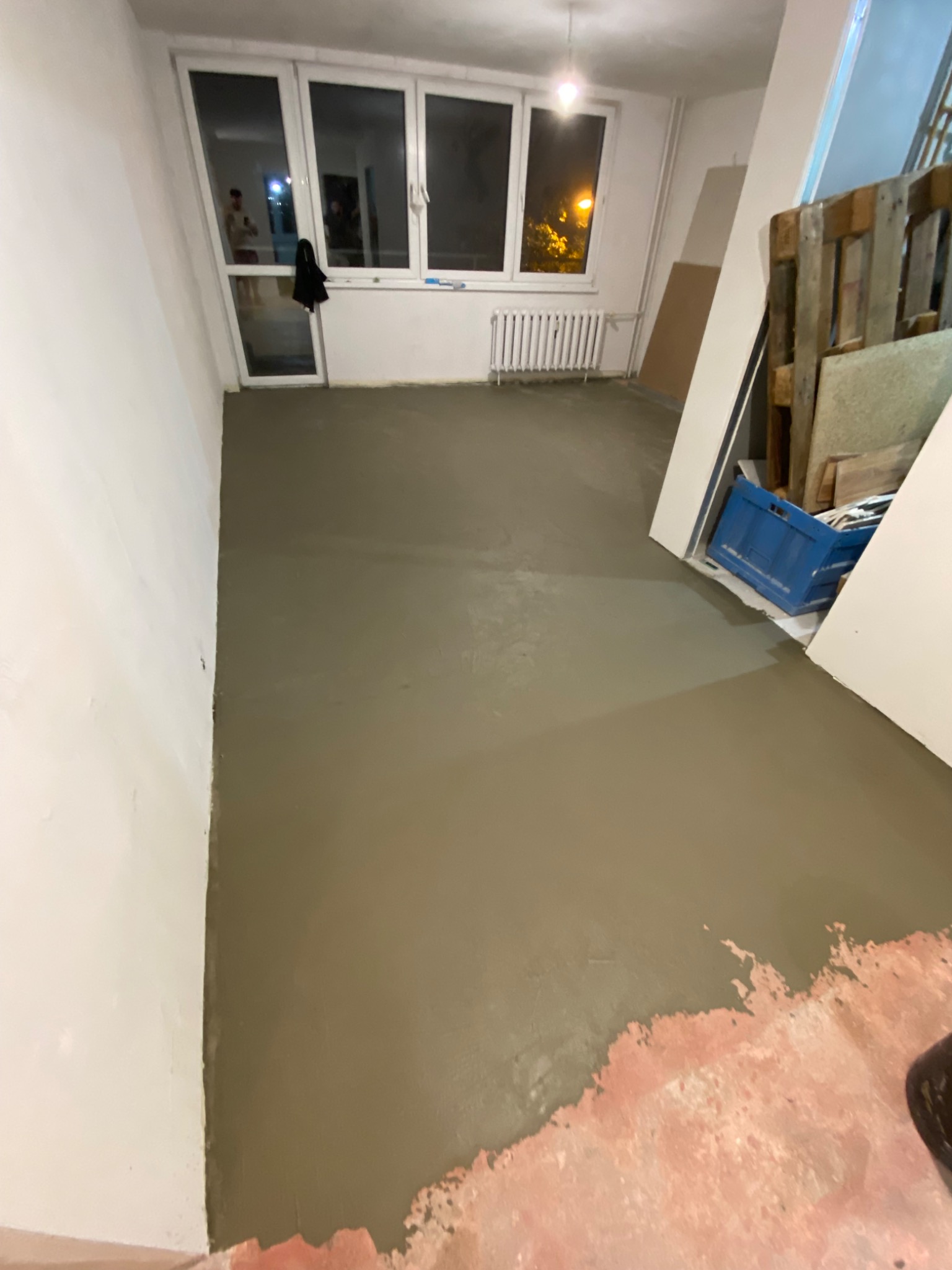 Wylewka samopoziomująca na podłodze w trakcie realizacji, widoczne ślady starej podłogi przy ścianie, okno z widokiem na noc, paleta drewniana i niebieski pojemnik w tle.