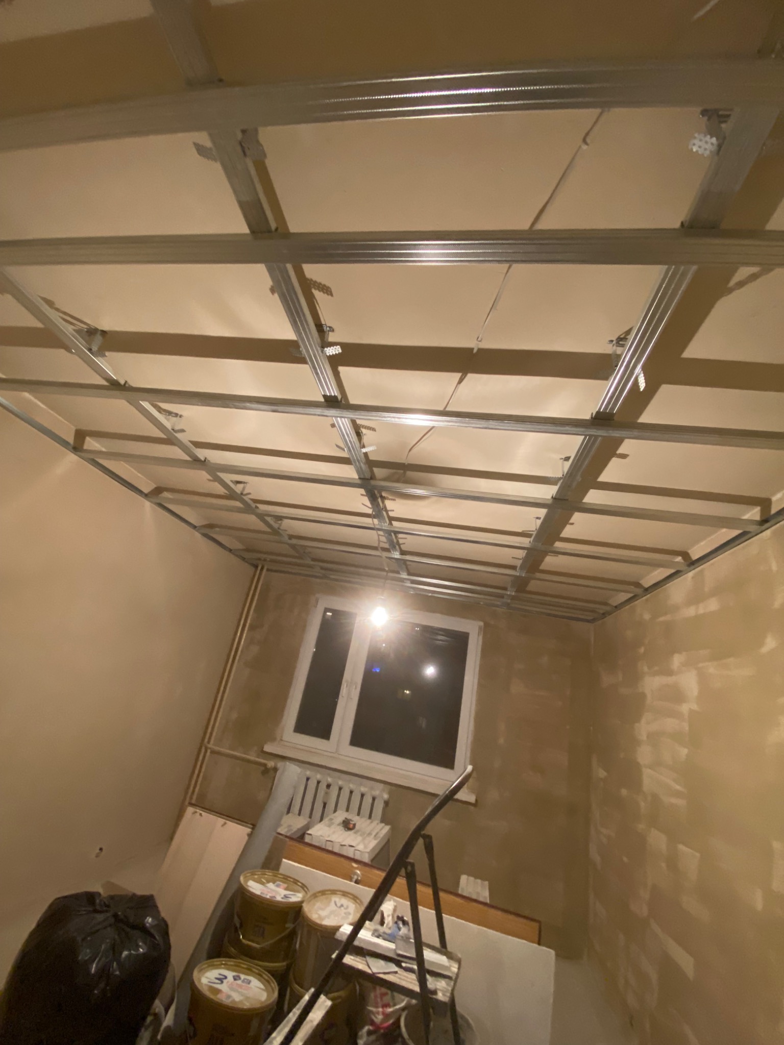 Montaż metalowej konstrukcji sufitu podwieszanego w trakcie remontu pokoju, widoczne profile i płyty gipsowe, ściany po wstępnym położeniu gładzi, okno z grzejnikiem, bałagan na podłodze.