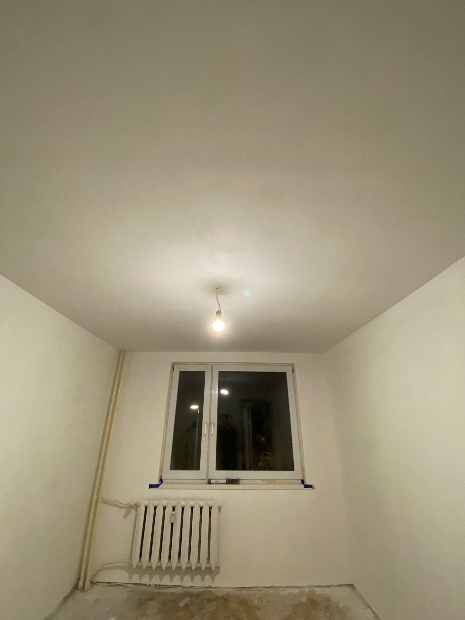 Wnętrze pokoju po malowaniu ścian i sufitu na biało, widoczny grzejnik, okno oraz goła żarówka zwisająca z sufitu.