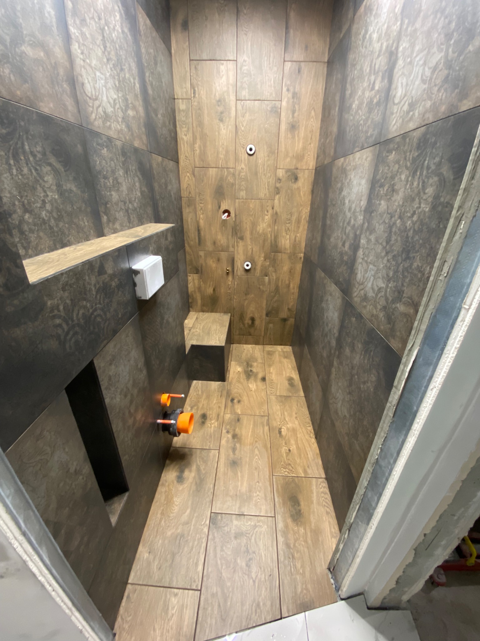Wnętrze kabiny prysznicowej w trakcie prac wykończeniowych, z widocznymi płytkami imitującymi drewno na podłodze i ścianach, instalacją wodną z pomarańczowymi elementami oraz półką wbudowaną w ścianę.