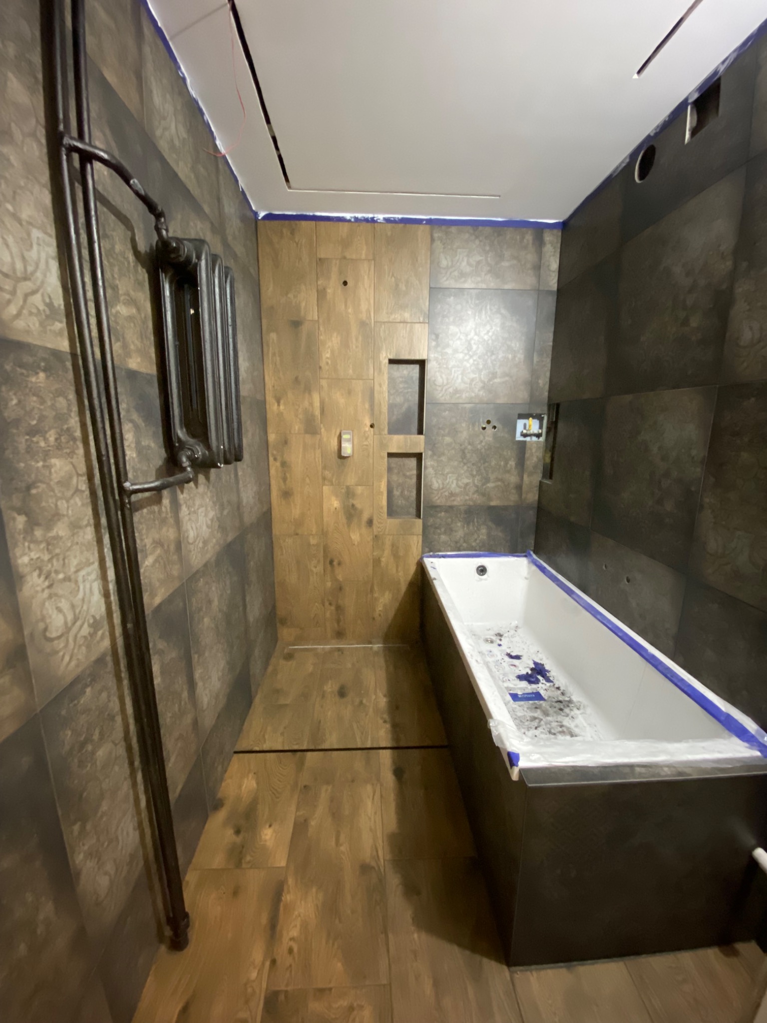 Łazienka w trakcie remontu: montaż płytek imitujących drewno na podłodze i ścianach, widoczna wanna zabezpieczona folią, grzejnik żeberkowy w stylu retro oraz przygotowane otwory pod armaturę.