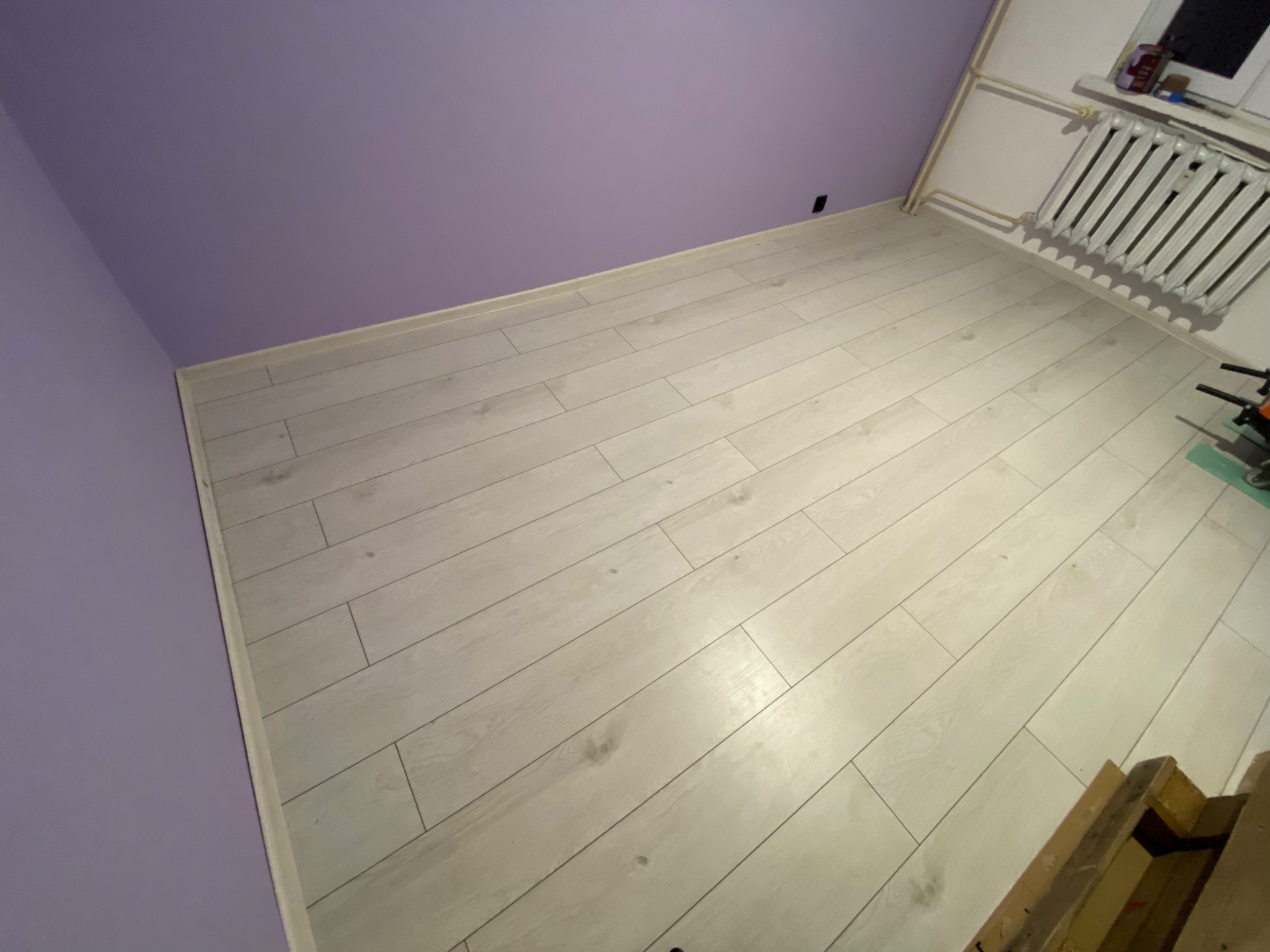 Jasne panele podłogowe ułożone w pokoju z fioletowymi ścianami, widoczny fragment grzejnika i okna w tle, oraz schodki w prawym dolnym rogu.