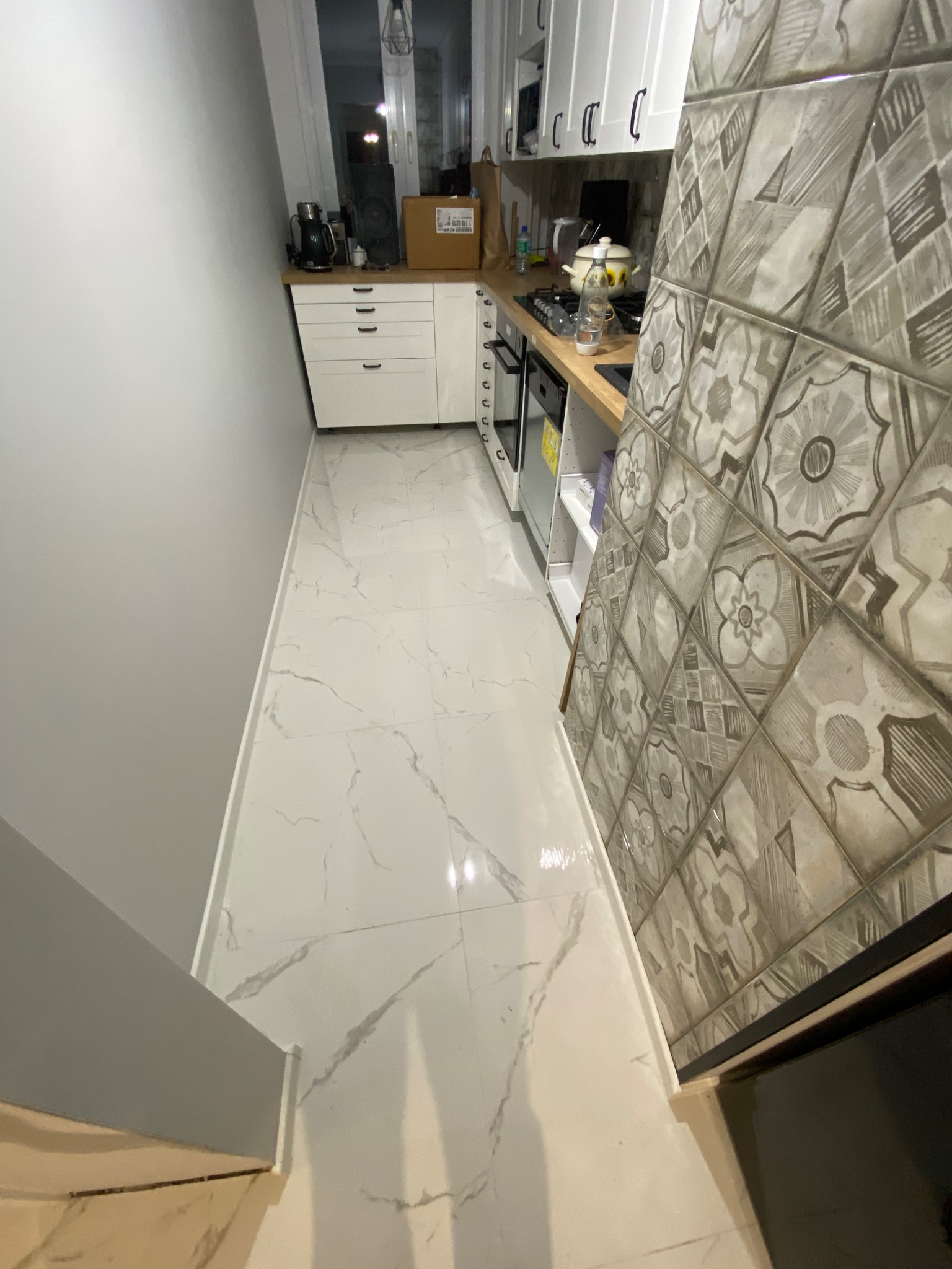 Wąska kuchnia z białymi szafkami, marmurową podłogą i ścianą wyłożoną dekoracyjnymi, wzorzystymi płytkami ceramicznymi w odcieniach szarości.