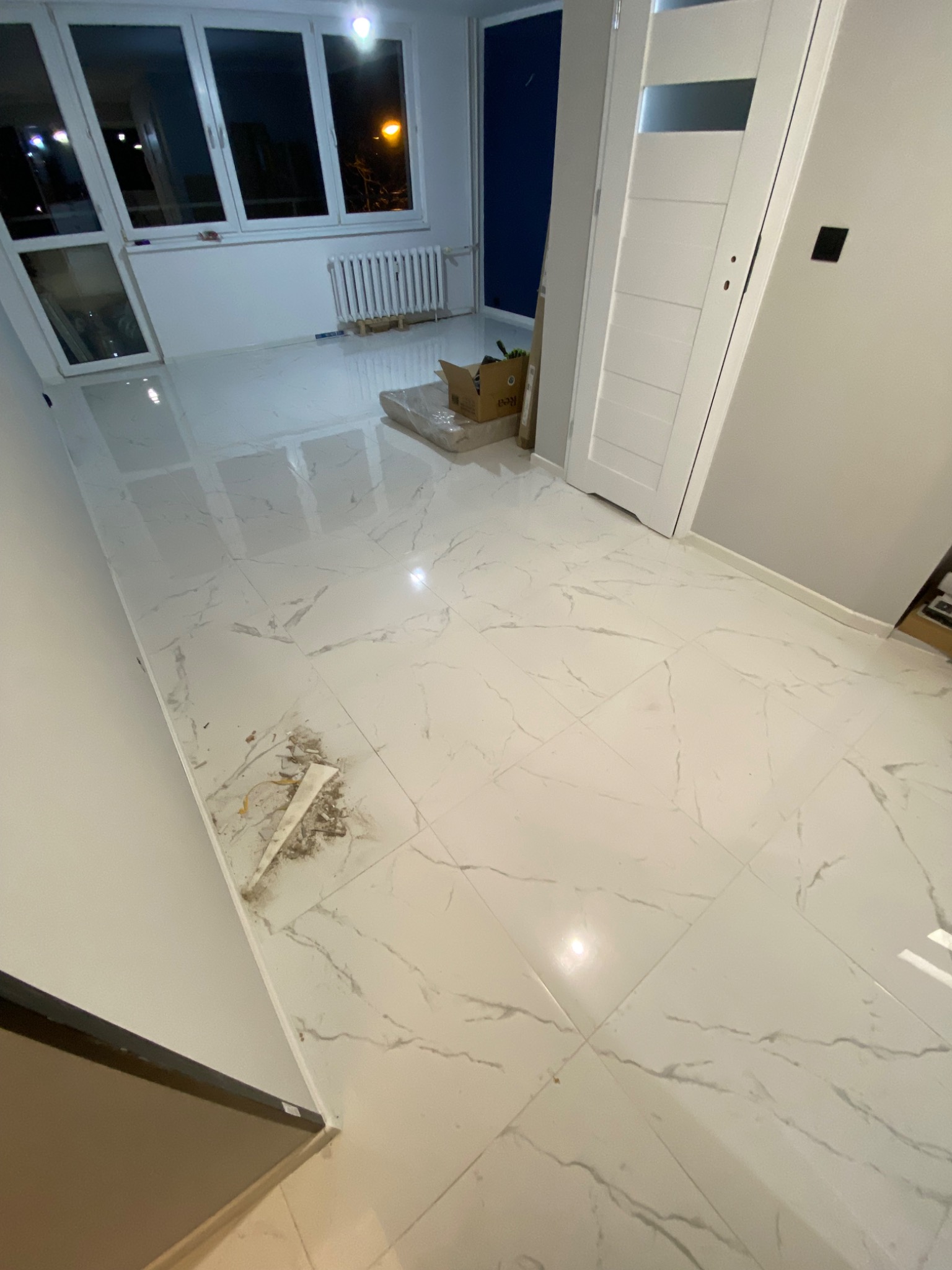 Białe, marmurkowe płytki podłogowe w trakcie układania w mieszkaniu w Mysłowicach, widoczne niedociągnięcia i resztki zaprawy, częściowo zakryte kartonem.