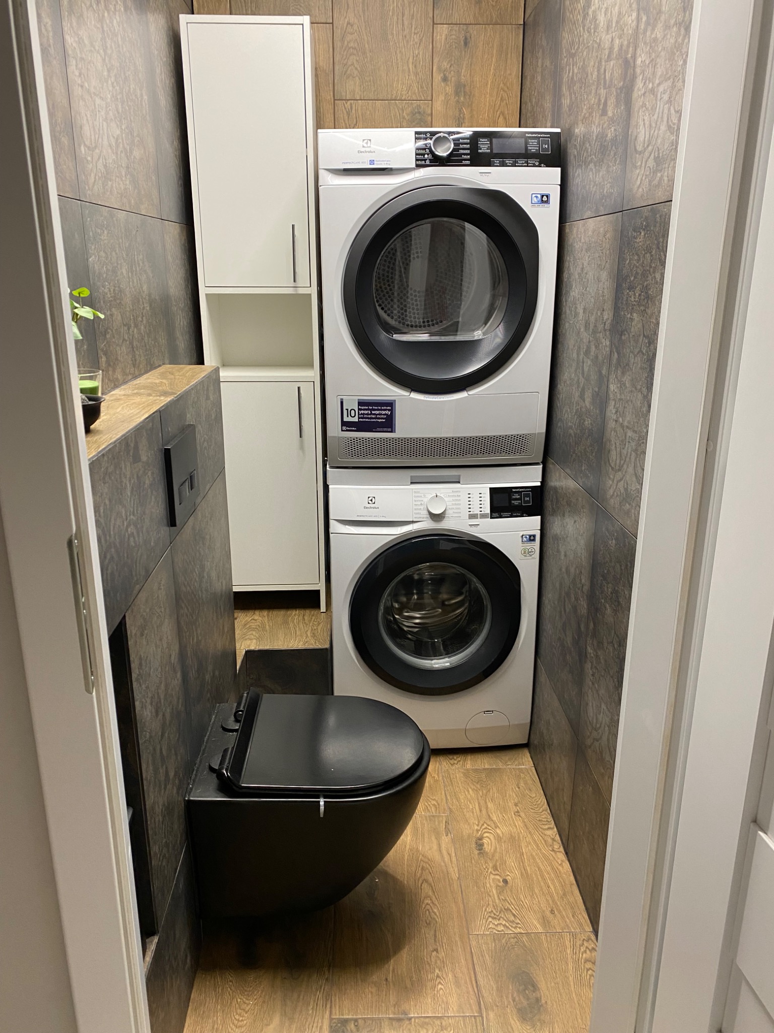 Nowoczesna łazienka z ciemnymi płytkami imitującymi drewno, podwieszaną toaletą w czarnym kolorze i pralko-suszarką Electrolux ustawioną w pionie, biała szafka w tle.