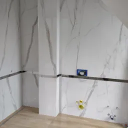 Łazienka w trakcie remontu, widoczne marmurowe płytki na ścianach z chromowanymi listwami, rury do podłączenia urządzeń sanitarnych i odpływ liniowy w podłodze wyłożonej panelami podłogowymi.