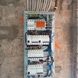 Otwarta skrzynka elektryczna z zainstalowanymi bezpiecznikami i przewodami na tle surowej ściany z cegły.